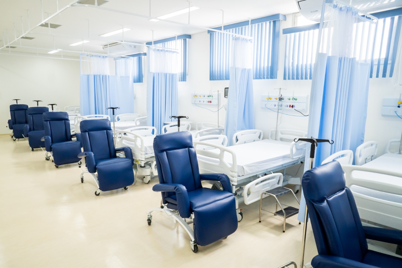 Foto de clínica - A grande sala branca conta com camas de UTI e equipamentos médicos na parede direita, ao pé de cada cama temos uma poltrona azul
